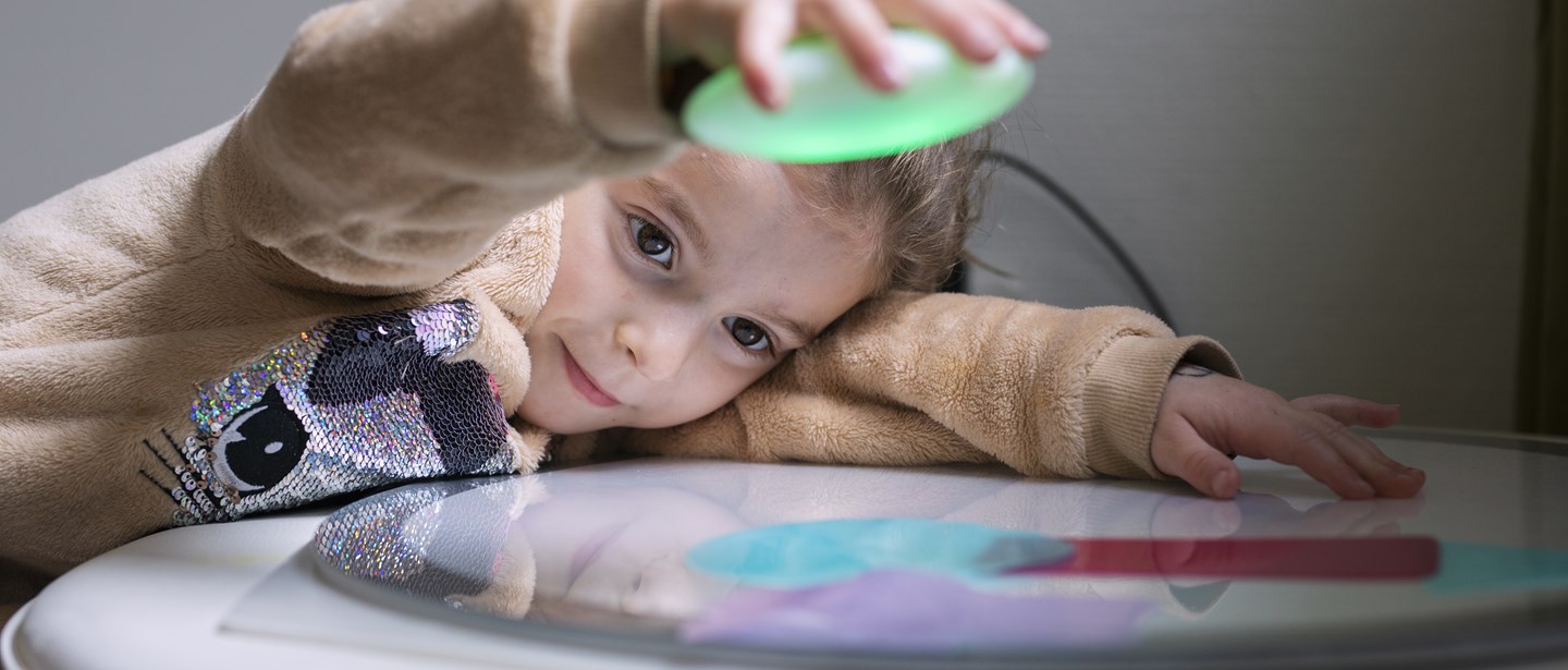 Ett foto av ett förskolebarn som liggandes mot en blank bordsyta lyfter en grön plastbit och tittar utforskande på reflektionen den ger.